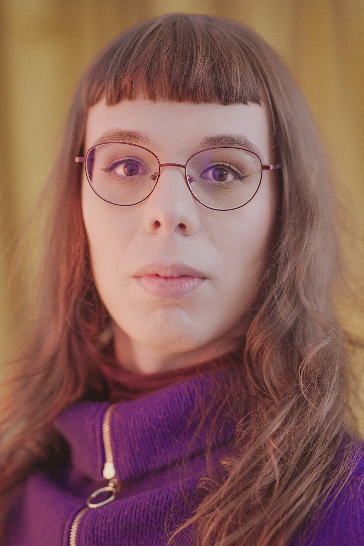 Portret kobiety w długich włosach i okularach, ubranej w fioletowy golf. W zamyśleniu i bez uśmiechu, spogląda wprost w obiektyw aparatu. 