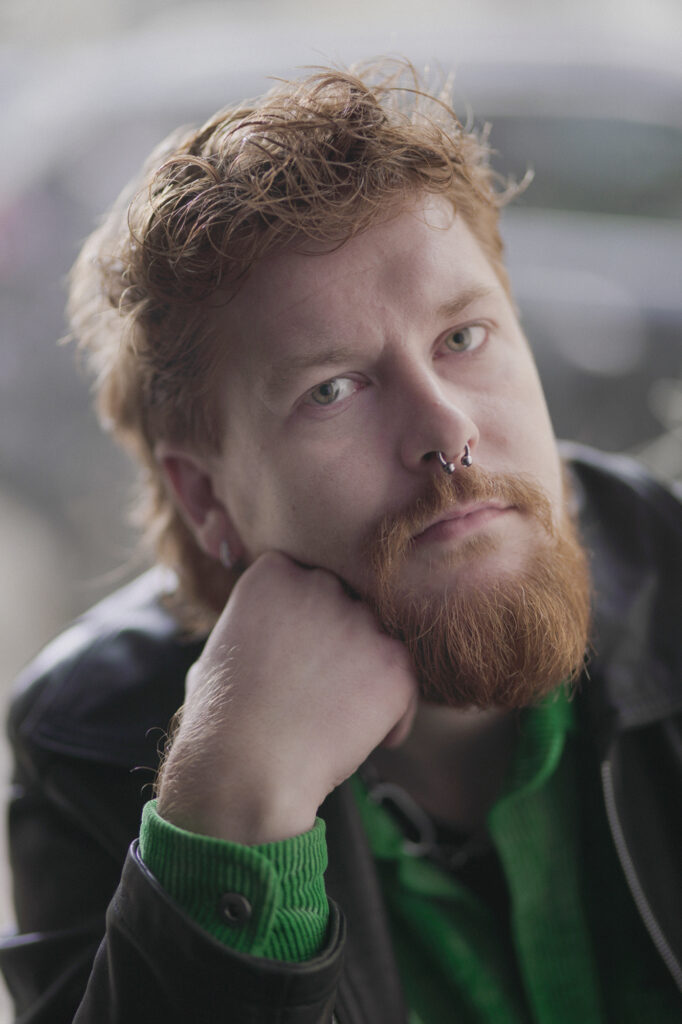 Mężczyzna ze zmierzwionymi włosami, z brodą i kolczykiem w nosie, podpiera twarz dłonią. Zamyślony, ze smutną miną, spogląda w obiektyw.